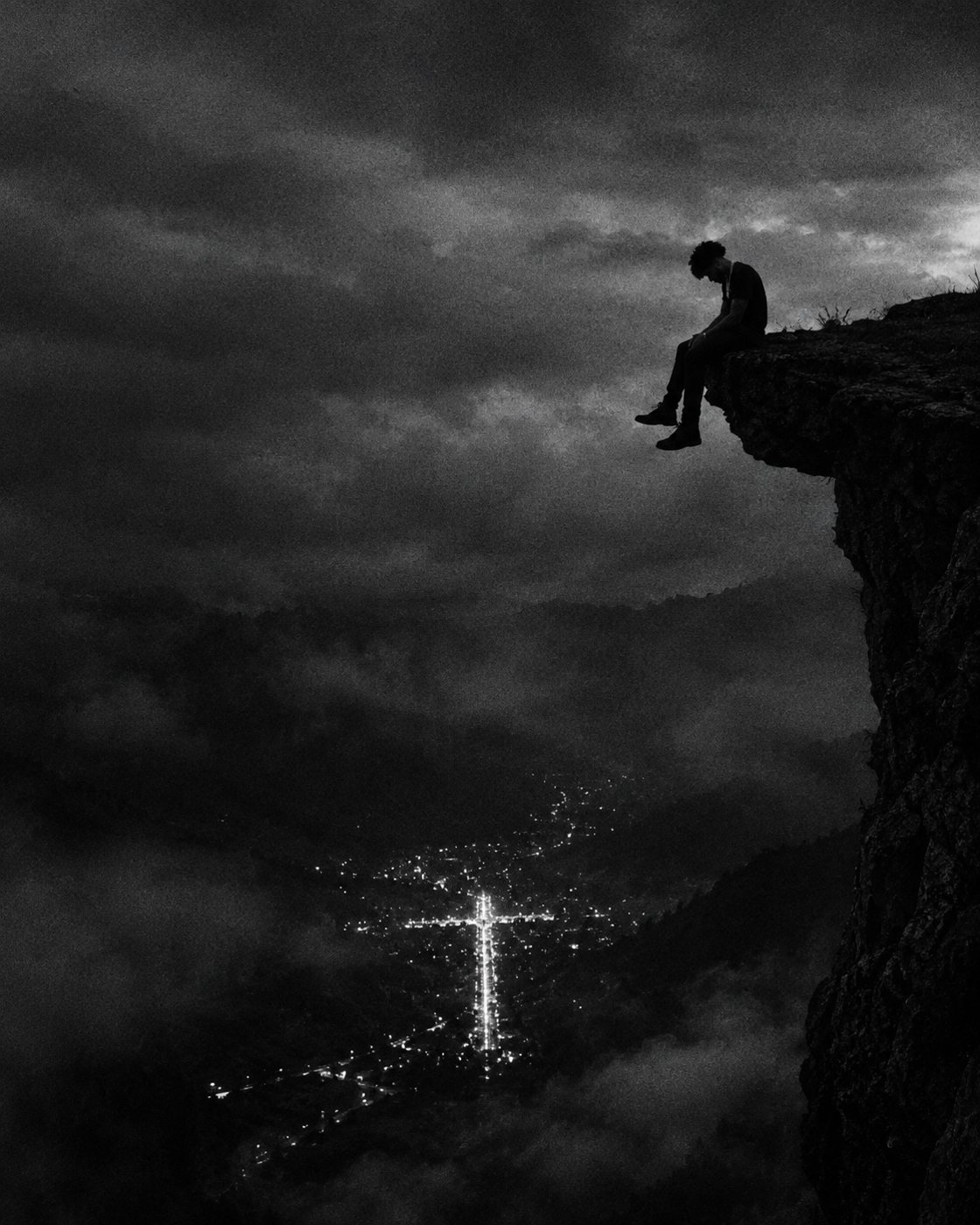 Figure sitting on a cliff edge above a foggy valley with city lights forming a cross